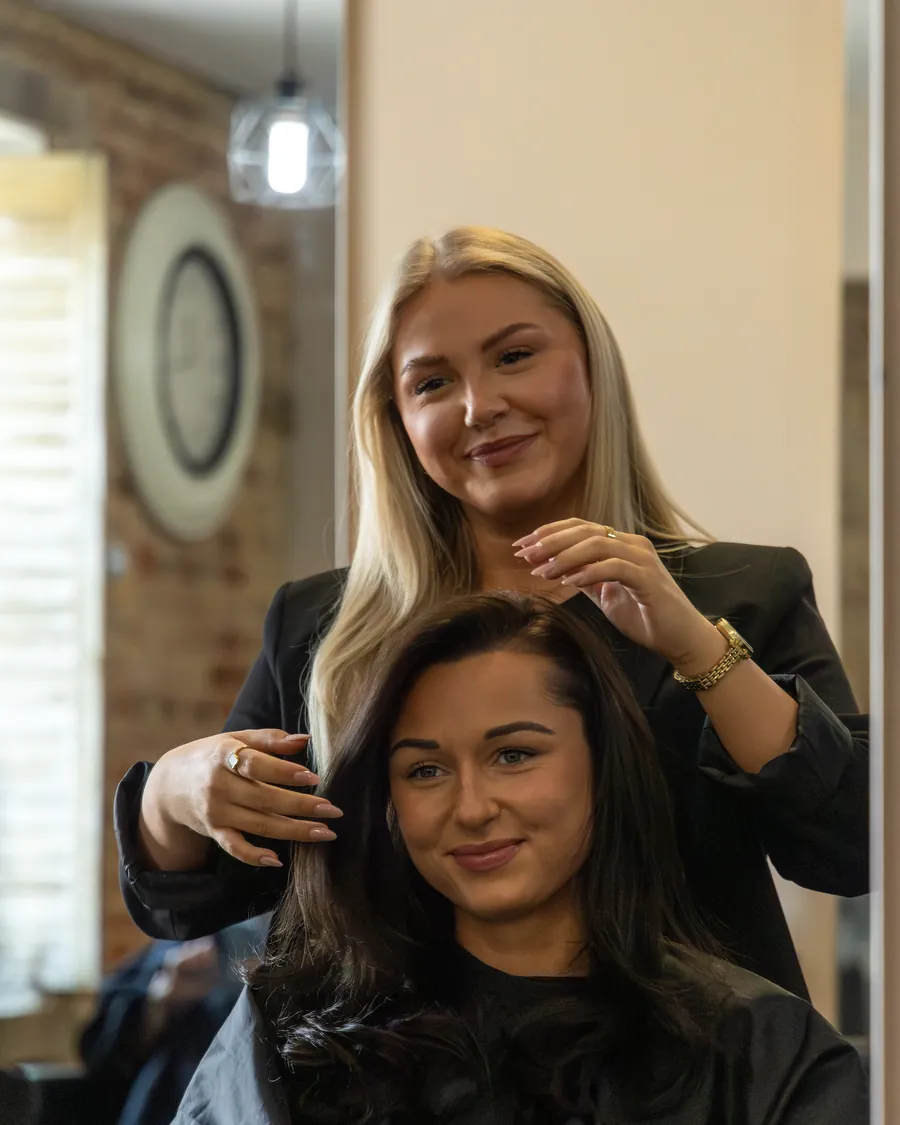 A hairdresser styling a client's long dark hair at the mirror