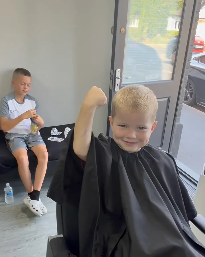 A young boy in a barber's cape flexing his arm after his haircut
